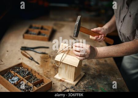 Closeup of hands making wooden birdhouse on workbench with tools, hammer and nails in workshop Stock Photo