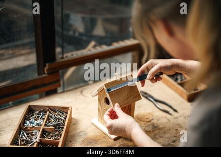 Female artisan measuring wood to make homemade birdhouse, surrounded by vintage tools and DIY equipment in workshop Stock Photo