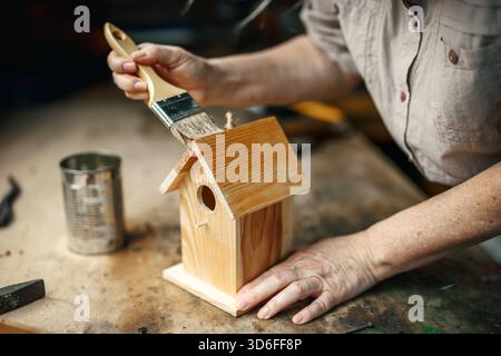 Woman painting protective wood stain to handmade birdhouse in workshop, ensuring durability for outdoor garden use Stock Photo