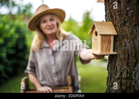 Smiling mature woman hanging birdhouse on tree in garden, supporting bird nesting and protection Stock Photo