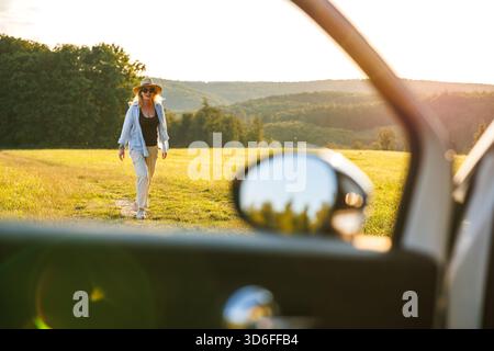 Happy smiling woman enjoying sunset during road trip stop. View through car window. Travel at summer vacation Stock Photo