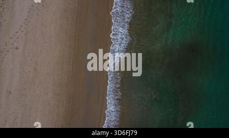 Sharp line of white waves breaking between the dark sand and the deep emerald-green ocean water Stock Photo