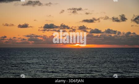 The sun sets over the horizon, casting a warm glow on the water and clouds above Stock Photo