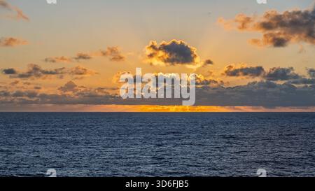 The sun sets over the horizon, casting a warm glow on the water and clouds above Stock Photo