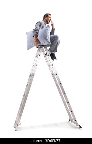 Man in pajamas with a pillow sitting on top of a ladder and yawning isolated on white background Stock Photo