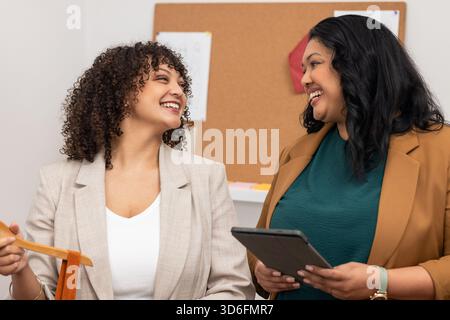 Two female colleagues exchanging ideas at office by cork board with fabric sample and tablet Stock Photo