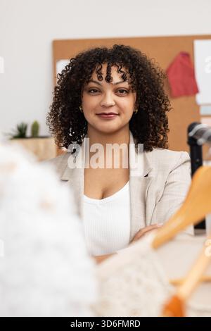 Hispanic female fashion designer sitting at desk in studio arranging hangers and draping garment Stock Photo