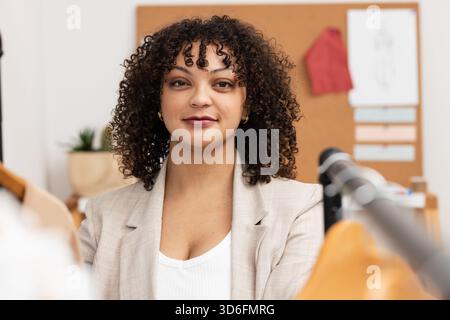 African American woman wearing blazer arranging garments on clothing rack in studio with cork board Stock Photo