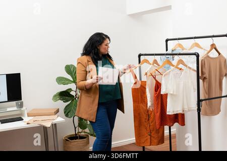 Asian woman holding paper while browsing black metal clothing rack with wooden hangers in office Stock Photo