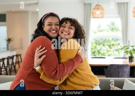 Hispanic mother hugging African American daughter at home on sofa near dining table, kitchen island Stock Photo