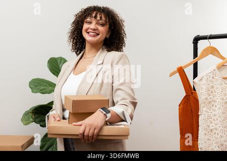 Woman wearing beige blazer carrying three cardboard boxes in boutique with clothing rack Stock Photo