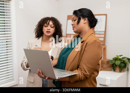 Diverse female coworkers standing at office examining silver laptop and smartphone by cork boards Stock Photo