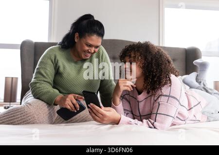 Mother and daughter pointing at smartphone screen while lying on bed in bedroom by windows Stock Photo