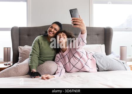 Mother and daughter sitting on bed in bedroom taking selfie with smartphones and smiling Stock Photo