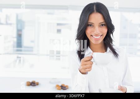 African American woman in shirt holding cup and saucer near pastries at office window, copy space. Breakroom, cityscape, blouse, bright, minimalist, p Stock Photo