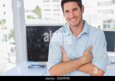 Large monitor displaying blurred icons, eyeglasses resting on desk near window in modern office. Portrait, professional, confident, smiling, workspace Stock Photo