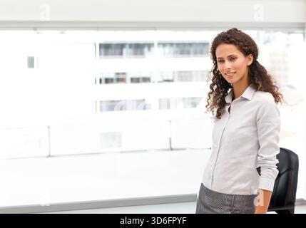 Adult woman leaning on black chair by large windows in light shirt, gray skirt, copy space. Office, modern, bright, minimal, urban, corporate, workspa Stock Photo