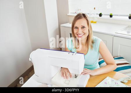 Female wearing striped top operating sewing machine at kitchen table, guiding fabric near plants. Stitching, bright, hobbyist, textile, daylight, work Stock Photo