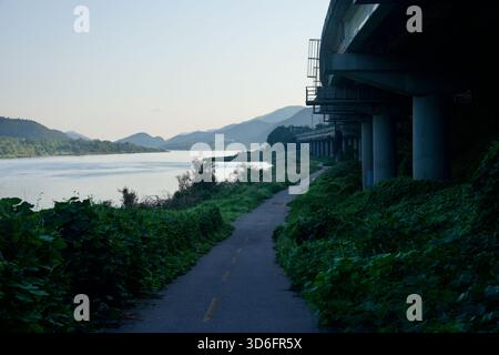 A quiet section of the Bukhangang Bicycle Path runs beneath an elevated expressway along the North Han River near Gapyeong, with layered hills fading Stock Photo