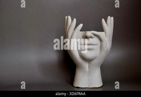 A plaster face sculpture framed by two hands, positioned against a neutral grey backdrop. Minimalist artistic still life with emotional symbolism. Stock Photo