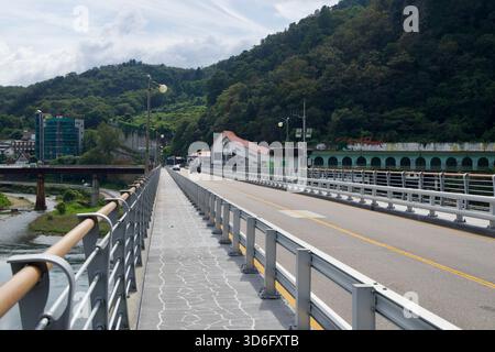 A pedestrian walkway runs along Gangchon Bridge with guardrails and lamp posts, while the river valley, village roofs and the long arcade on the oppos Stock Photo