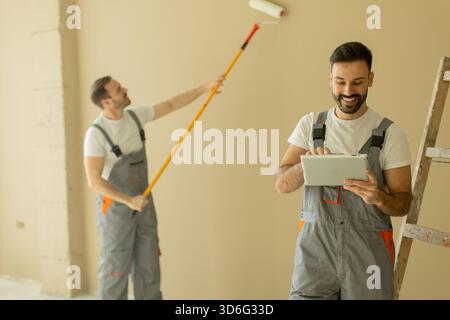 Two men in work overalls are repainting an interior wall. One is using a roller to apply paint, while the other checks tasks on a tablet in a renovate Stock Photo