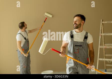Two men are engaged in painting a room with paint rollers. They wear overalls and are focused on transforming the wall color. Tools and a ladder can b Stock Photo