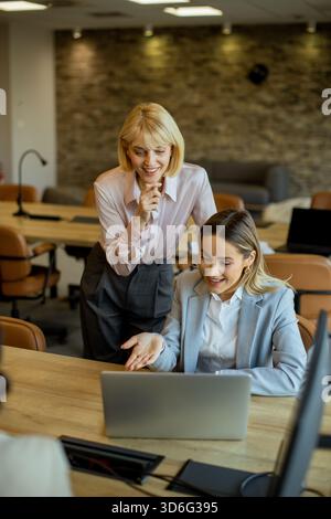 Two women are engaged in an animated discussion at a wooden table in a modern office. One points to the laptop screen while the other smiles and liste Stock Photo
