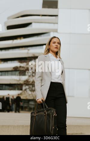 A young woman in a tailored suit is walking confidently in a modern urban setting. She carries a black bag and looks determined, showcasing a professi Stock Photo
