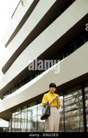 A woman dressed in a bright yellow blouse stands outside a contemporary building, smiling while looking at her smartphone. The sleek, curved design of Stock Photo