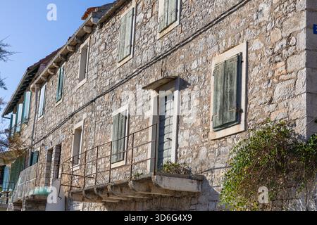 A weathered stone building with green wooden shutters, a small balcony, and iron railing along a sunlit street. The rustic texture and aged charm evok Stock Photo