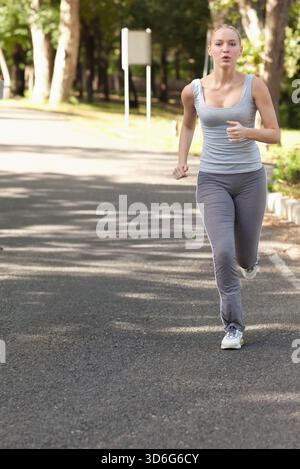 Female runner running along paved tree-lined road in park wearing gray tank and white running shoes. Outdoor, nature, athlete, path, sunlight, focus, Stock Photo