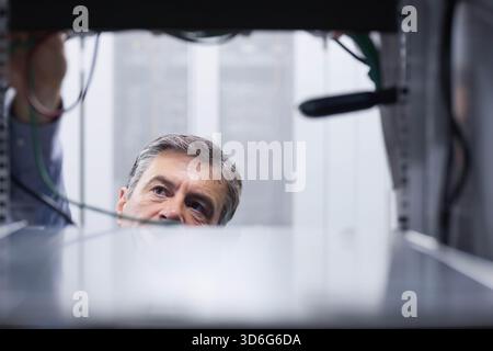 Mature adult male technician leaning into server rack, inspecting bundled cables and components. Cabinet, datacenter, cabling, engineering, inspection Stock Photo