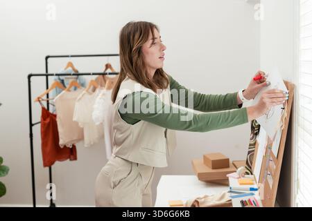 Woman arranging paper design sketches on corkboard in studio with clothing rack and tape Stock Photo