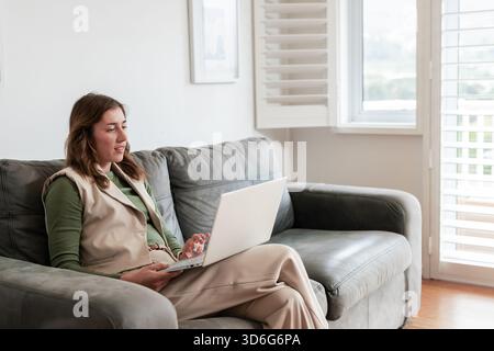 Woman in thirties sitting at laptop on grey couch at home wearing work attire, copy space Stock Photo