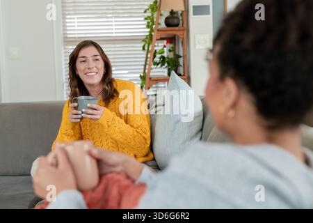 Diverse female friends chatting, holding grey and pink mugs on couch in living room, copy space Stock Photo