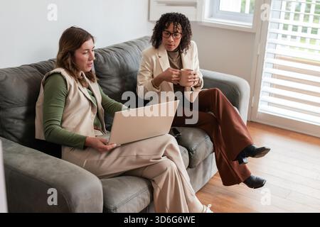 Women colleagues sitting on couch navigating laptop holding coffee mug with smartphone between them Stock Photo