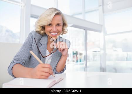 Mid-aged woman in gray blazer holding tortoise specs writing with pencil on white desk by windows. Office, modern, sunlit, minimal, notebook, glasses, Stock Photo