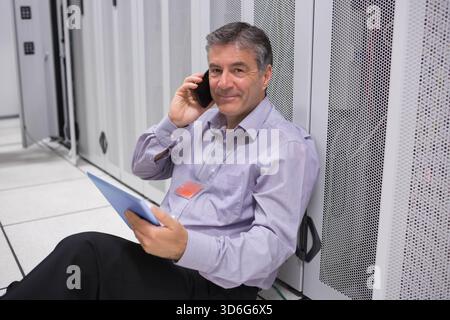 Senior man sitting on raised floor in data center, holding blue tablet and talking on smartphone. Server, cabinet, rack, technician, badge, lanyard, c Stock Photo