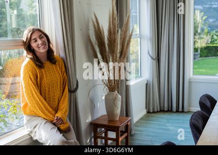 Woman wearing sweater leaning against window sill in dining area with pampas grass vase, copy space Stock Photo