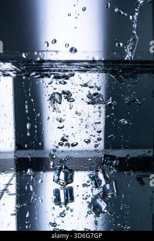8.Ice cubes are dropped into a transparent water tank, creating visible splashes, suspended droplets,rising bubbles. Blue lighting highlights fluid mo Stock Photo
