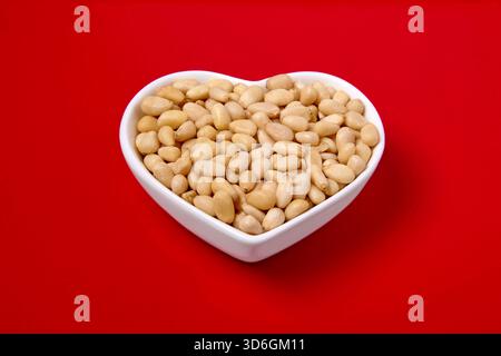 peeled pine nuts in a white heart-shaped bowl on a red background. Stock Photo