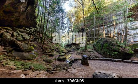 View of sunlit trees towering over moss-covered rocks and a small stream, creating a serene and inviting trail, Logan, Ohio, United States. Stock Photo