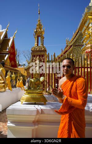 Young Buddhist monk clasping a lotus blossom in praying hands wearing a orange robe and sunglasses in front of golden statues at Wat Doi Suthep Stock Photo