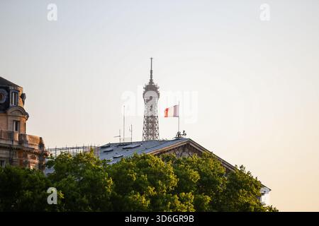 View of the Eiffel Tower rising behind rooftops and a French flag at sunrise in Paris, France. Stock Photo