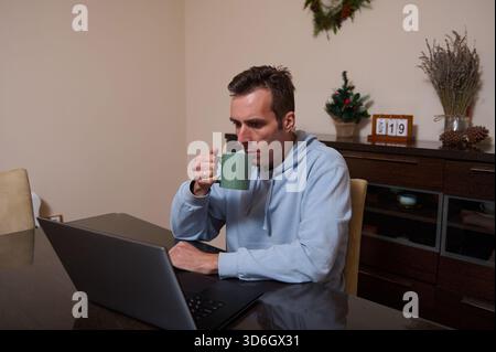 A man in a light blue hoodie works remotely at a dining table, sipping coffee from a mug. Christmas decor and a calendar setup create a warm home-offi Stock Photo
