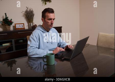 A man in a light blue hoodie works on a laptop at a home office, sipping coffee from a mug and concentrating on his tasks. Stock Photo