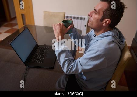 A man in a blue hoodie sits at a dining table with a laptop, sipping coffee from a mug. He works remotely from home, relaxed and focused. Stock Photo