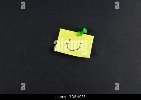 Yellow sticky note with smiley face pinned by green pushpin on black background, conveying positivity through simple, bold contrast. Stock Photo