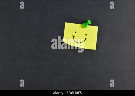 Yellow sticky note with smiley face pinned by green pushpin on black background, conveying positivity through simple, bold contrast. Stock Photo